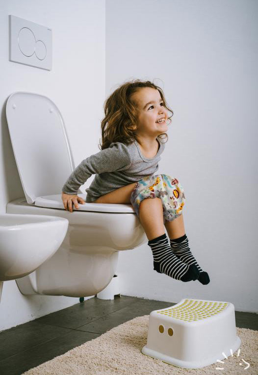 Χαμογελαστό κοριτσάκι κάθεται στην τουαλέτα και φοράει γκρι μπλούζα και Βρακάκι Εκμάθησης Μωρού Popolini με ζωάκια και μπλε άσπρο ριγέ κάλτσες από οργανικό βαμβάκι