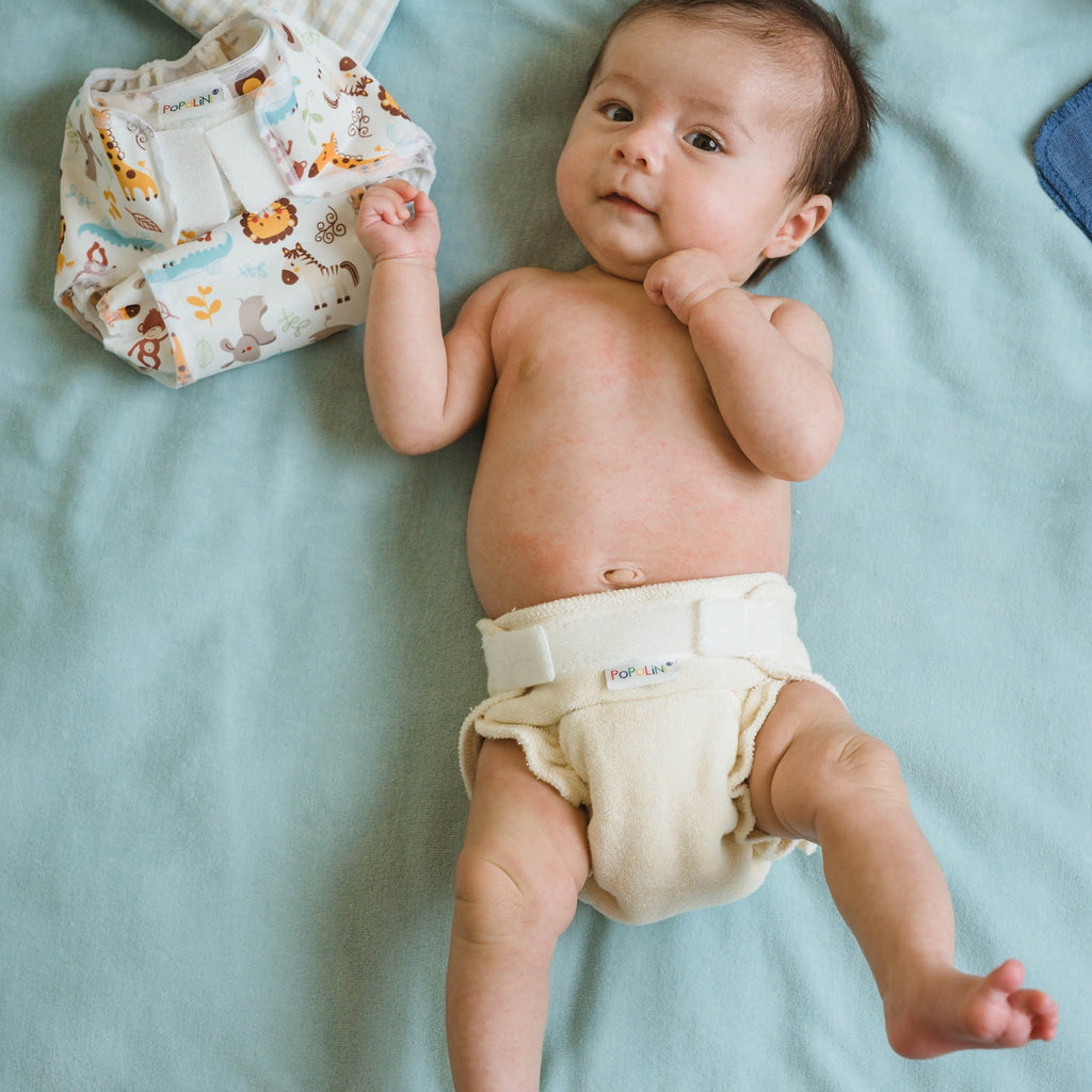 Νεογέννητο μωρό ξαπλωμένο σε σελτεδάκι γαλάζιο, φοράει υφασμάτινη πάνα για νεογέννητο Minifit Popolini σε φυσικό χρώμα. Δίπλα στο μωρό απεικονίζεται ένα βρακάκι πάνας Popolini με σχέδιο ζώα της ζούγκλας. 