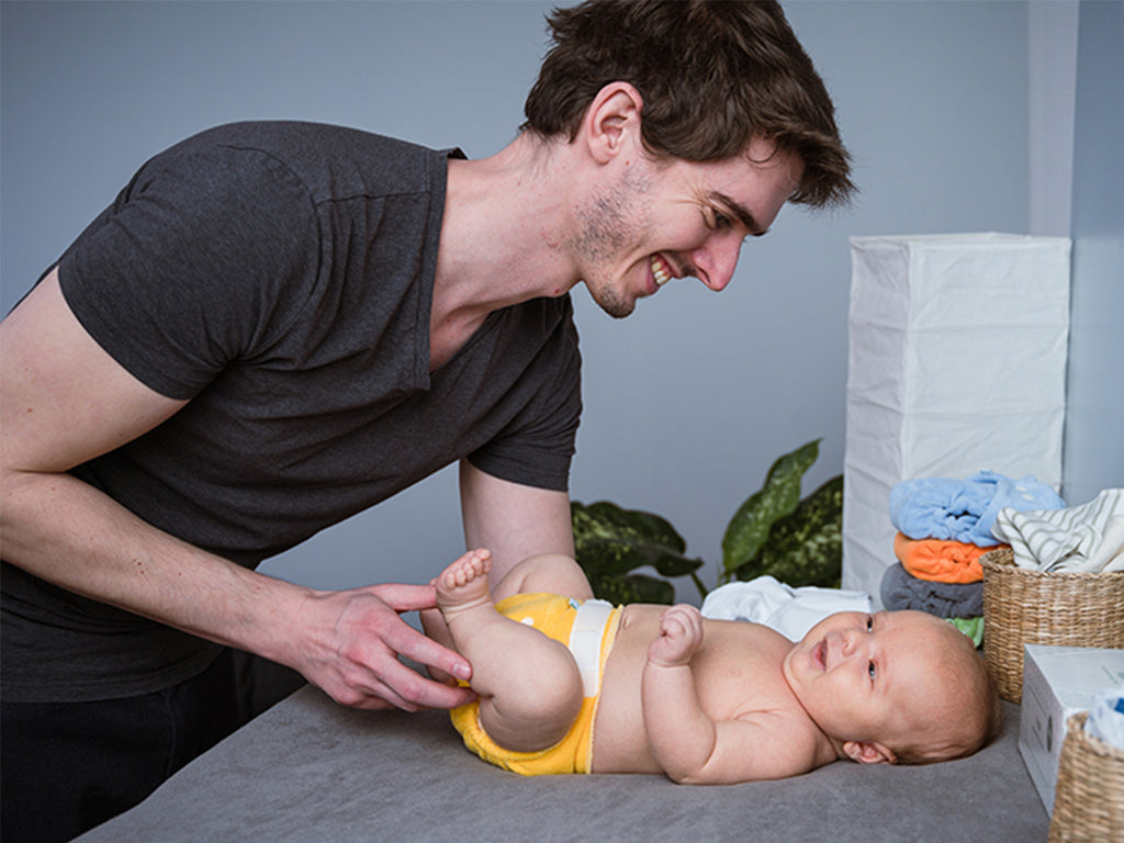 Μπαμπάς χαμογελάει και αλλάζει πάνα στο μωρό. Το μωρό είναι ξαπλωμένο στην αλλαξιέρα και φοράει υφασμάτινη πάνα για νεογέννητο Minifit Popolini από πετσετέ ύφασμα σε κίτρινο χρώμα.