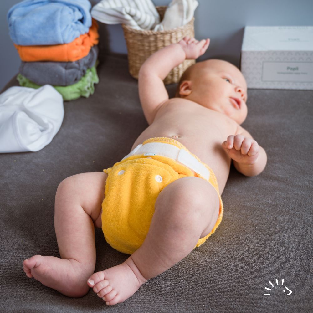 Νεογέννητο μωρό ξαπλωμένο σε γκρι σελτεδάκι, φοράει υφασμάτινη πάνα για νεογέννητο Minifit Popolini σε κίτρινο χρώμα. Δίπλα στο μωρό απεικονίζονται μουσελίνες για μωρό, ένθετα Popli και υφασμάτινες πάνες σε διάφορα χρώματα. 